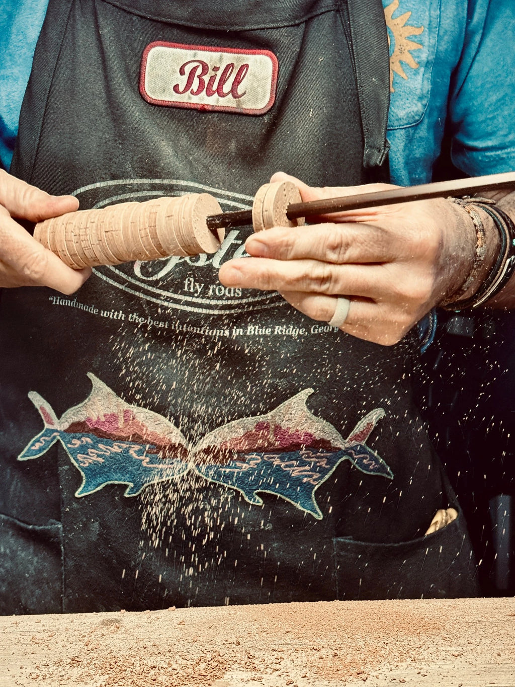 Craftsman shaping a cork grip by hand on a bamboo fly rod blank, with cork dust flying.